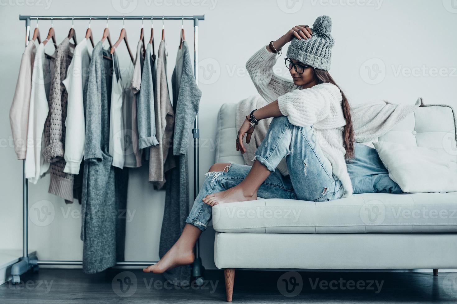 Choosing the right option.  Thoughtful young woman in casual wear adjusting her hat and looking away while sitting on the couch at home near her clothes hanging on the racks photo