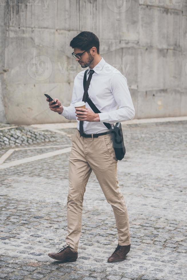 Sending quick business message. Full length of confident young man in glasses holding coffee cup and using his smart phone while walking outdoors photo