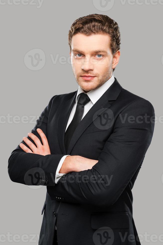 Successful businessman. Portrait of confident young man in formalwear looking at camera while keeping arms crossed and standing against grey background photo