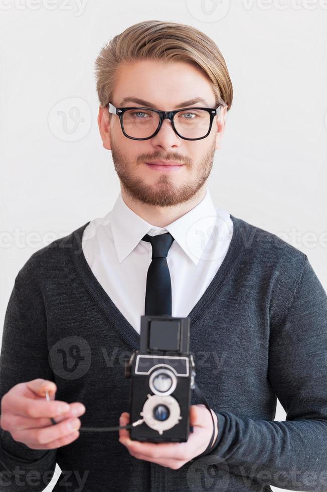 fotógrafo de estilo antiguo. un joven apuesto con anteojos sosteniendo una cámara retro mientras se enfrenta a un fondo gris foto