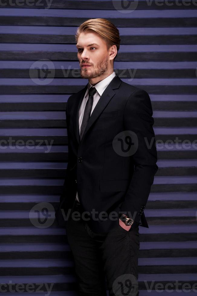 Thoughtful handsome. Side view of handsome young man in formalwear holding hands in pockets and looking away while standing against striped background photo