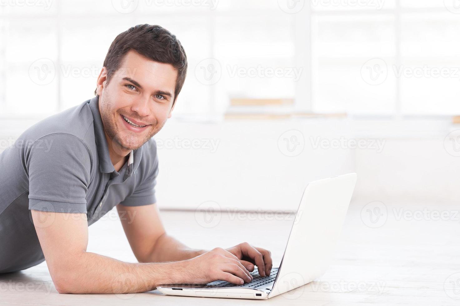 Surfing web at home. Cheerful young man lying on the floor with laptop and looking at camera photo