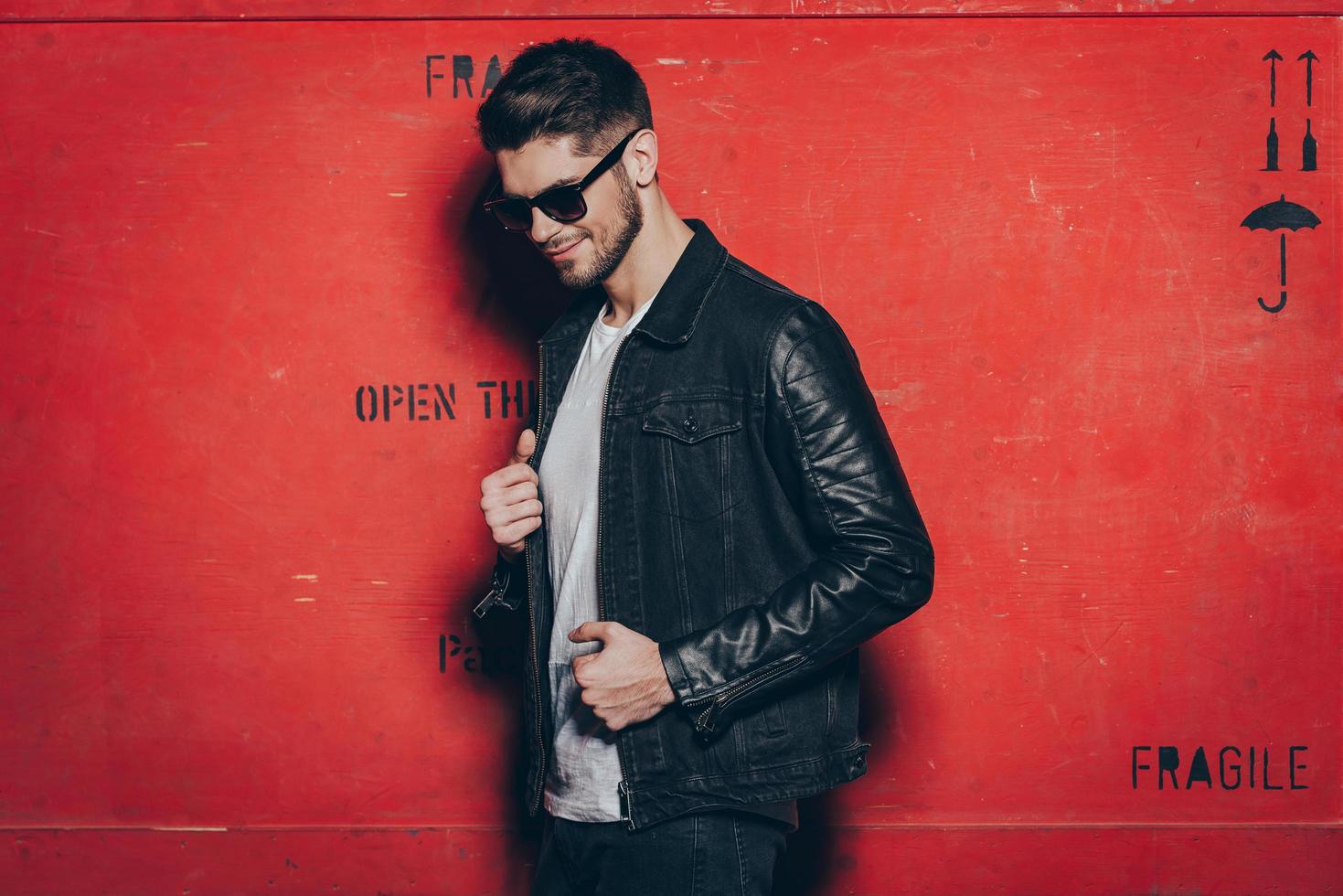 mirada de moda un joven apuesto con gafas de sol ajustando su chaqueta y mirando hacia otro lado con una sonrisa mientras se enfrenta a un fondo rojo foto