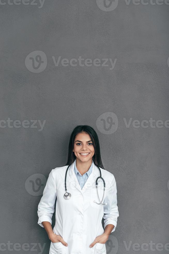 Confident female doctor. Confident young female doctor in white lab coat holding hands in pockets and smiling while standing against grey background photo