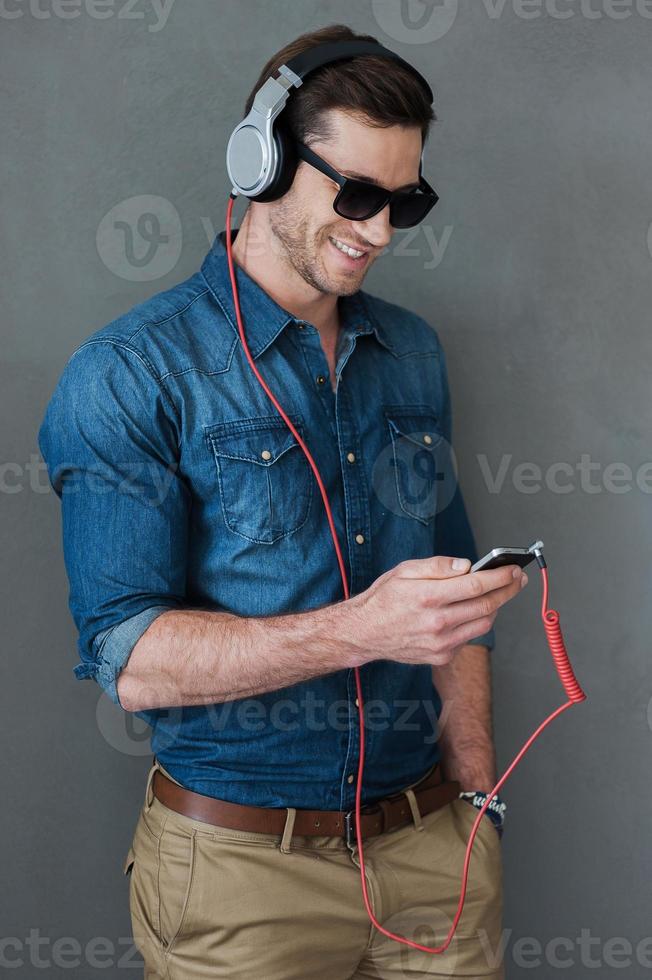 disfrutando de la música favorita. un joven alegre con auriculares sosteniendo un reproductor de mp3 y mirándolo mientras se enfrenta a un fondo gris foto