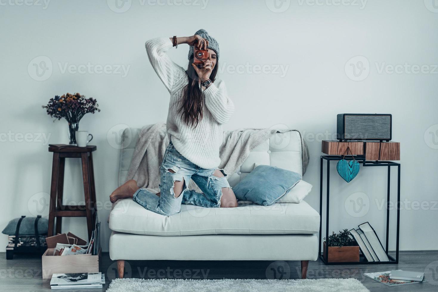 Smile  Beautiful young woman in casual wear and knit hat taking a photo by her retro camera while standing on the sofa at home