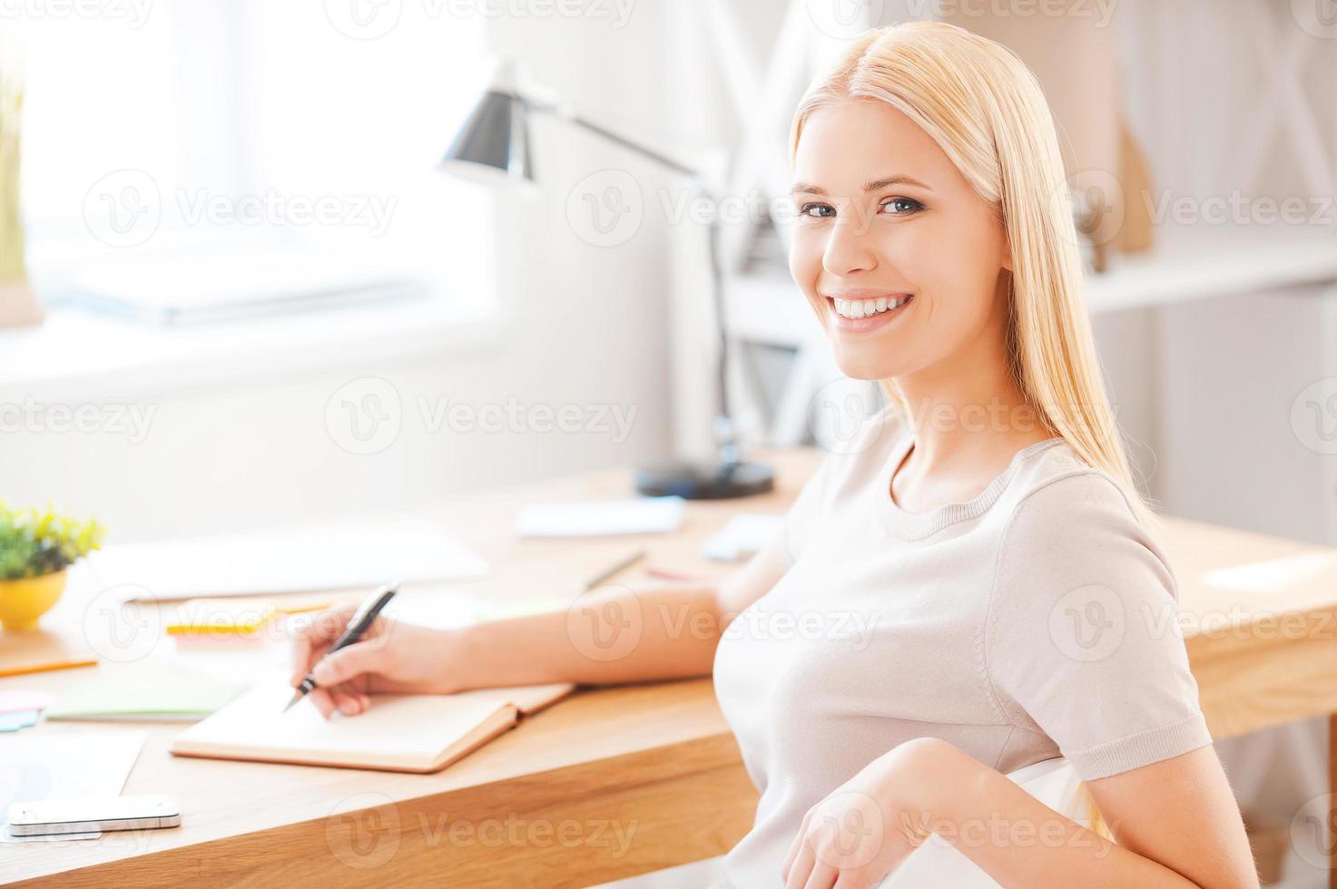 trabajando con placer. bella joven escribiendo algo en un bloc de notas y sonriendo mientras se sienta en su lugar de trabajo en la oficina foto