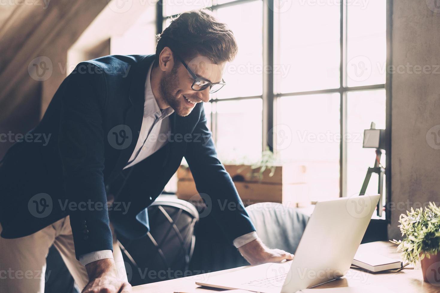 Best solutions every day. Confident young man in smart casual wear looking at laptop and smiling while standing near his working place in office photo