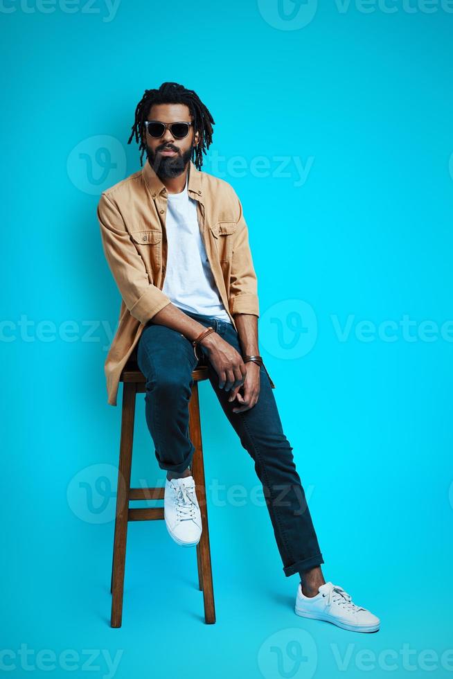 Full length of charming young African man in casual wear looking at camera while sitting on stool against blue background photo