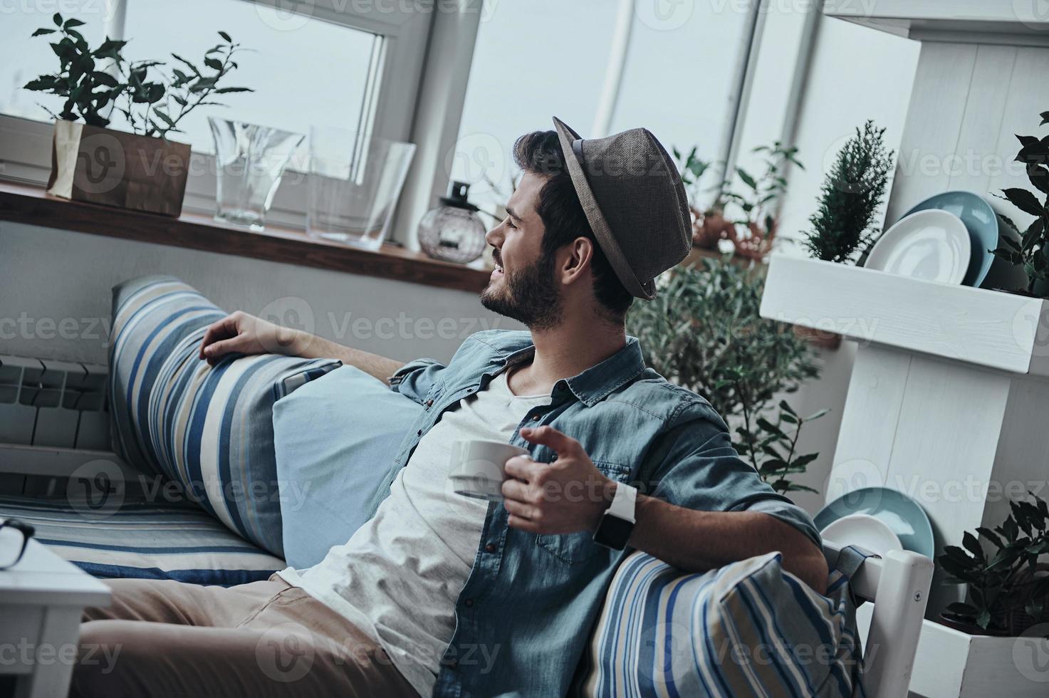 Enjoying fresh coffee. Thoughtful young man holding a cup and looking outside while sitting on the sofa photo