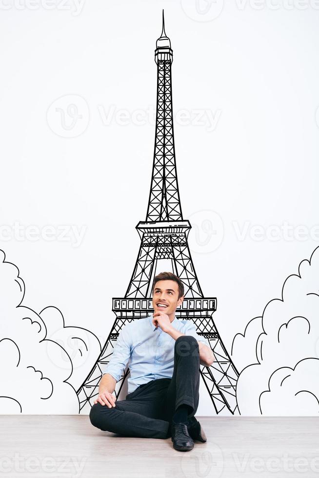 Dreaming about Paris. Young handsome man keeping hand on chin and looking away with smile while sitting on the floor with illustration of Eiffel Tower at the background photo