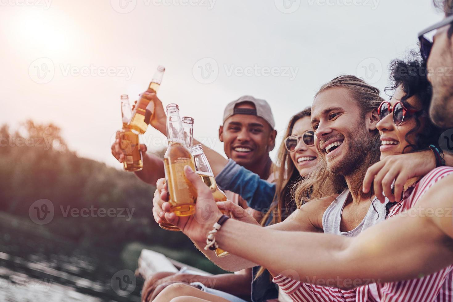 Just relaxing. Group of happy young people in casual wear smiling and toasting each other while sitting on the pier photo
