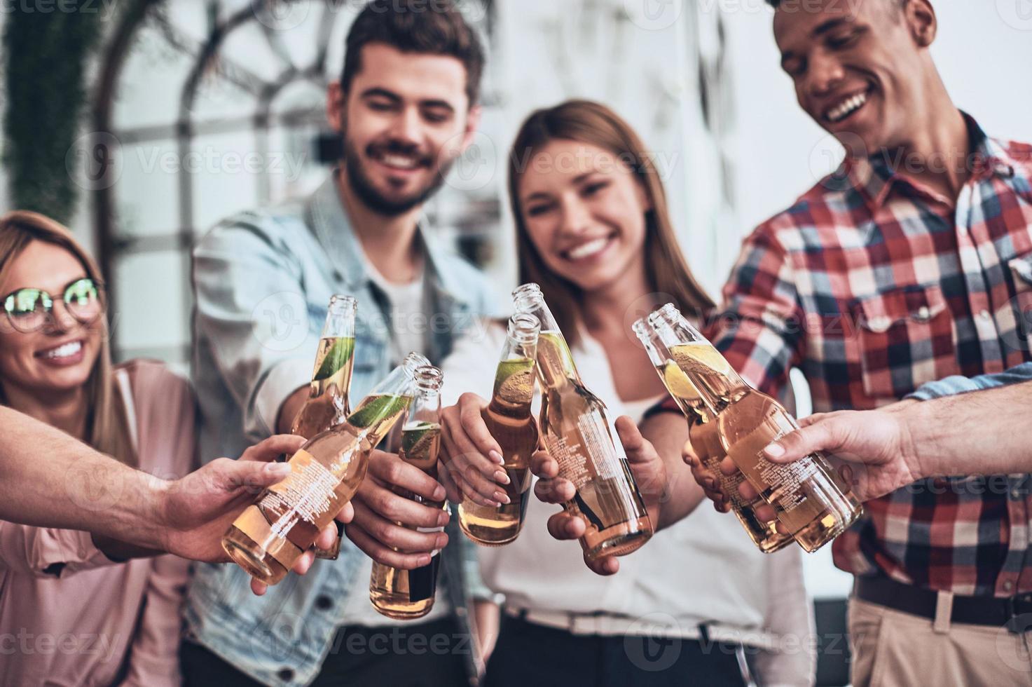 To us.  Group of young people in casual wear toasting each other and smiling while having a party indoors photo