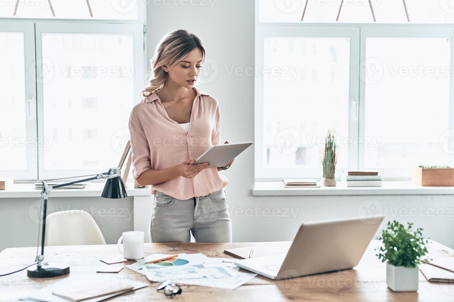 New decisions every day. Beautiful young woman in smart casual wear working using digital tablet while standing in modern office photo
