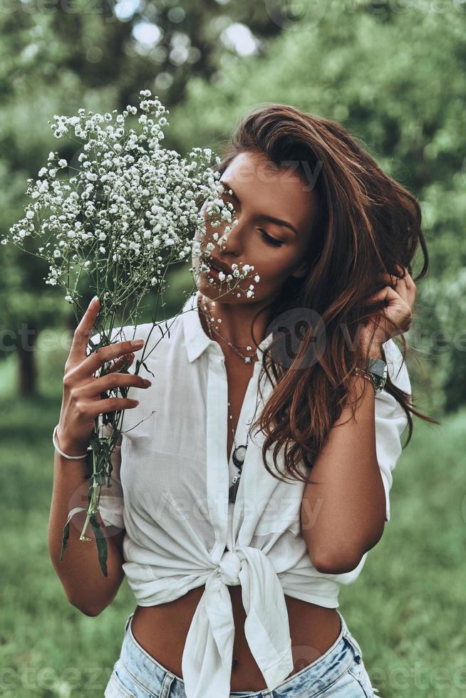 disfrutando de la primavera. mujer joven atractiva en ropa casual sosteniendo flores y manteniendo la mano en el cabello mientras está de pie al aire libre foto