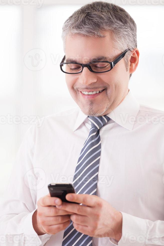 Typing a business message. Cheerful senior man in shirt and tie holding a mobile phone and smiling photo