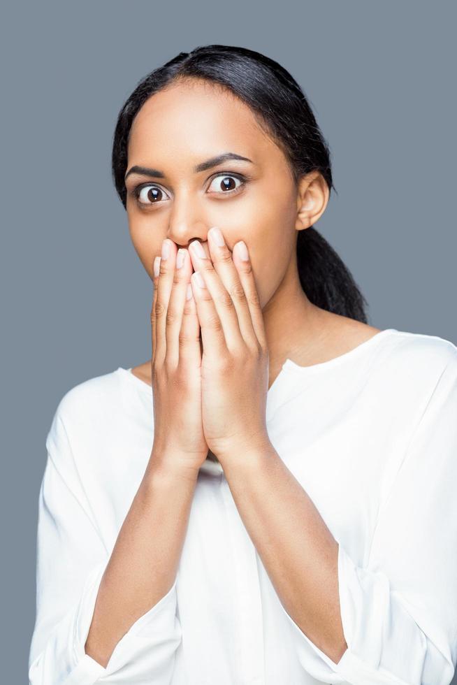 Shocking news. Shocked young African woman covering mouth with hands while standing against grey background photo