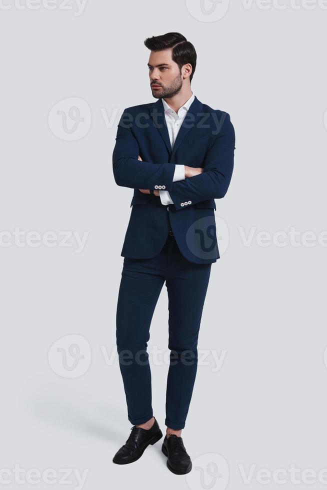 Perfect in his style. Full length of handsome young man in full suit looking away and keeping arms crossed while standing against grey background photo