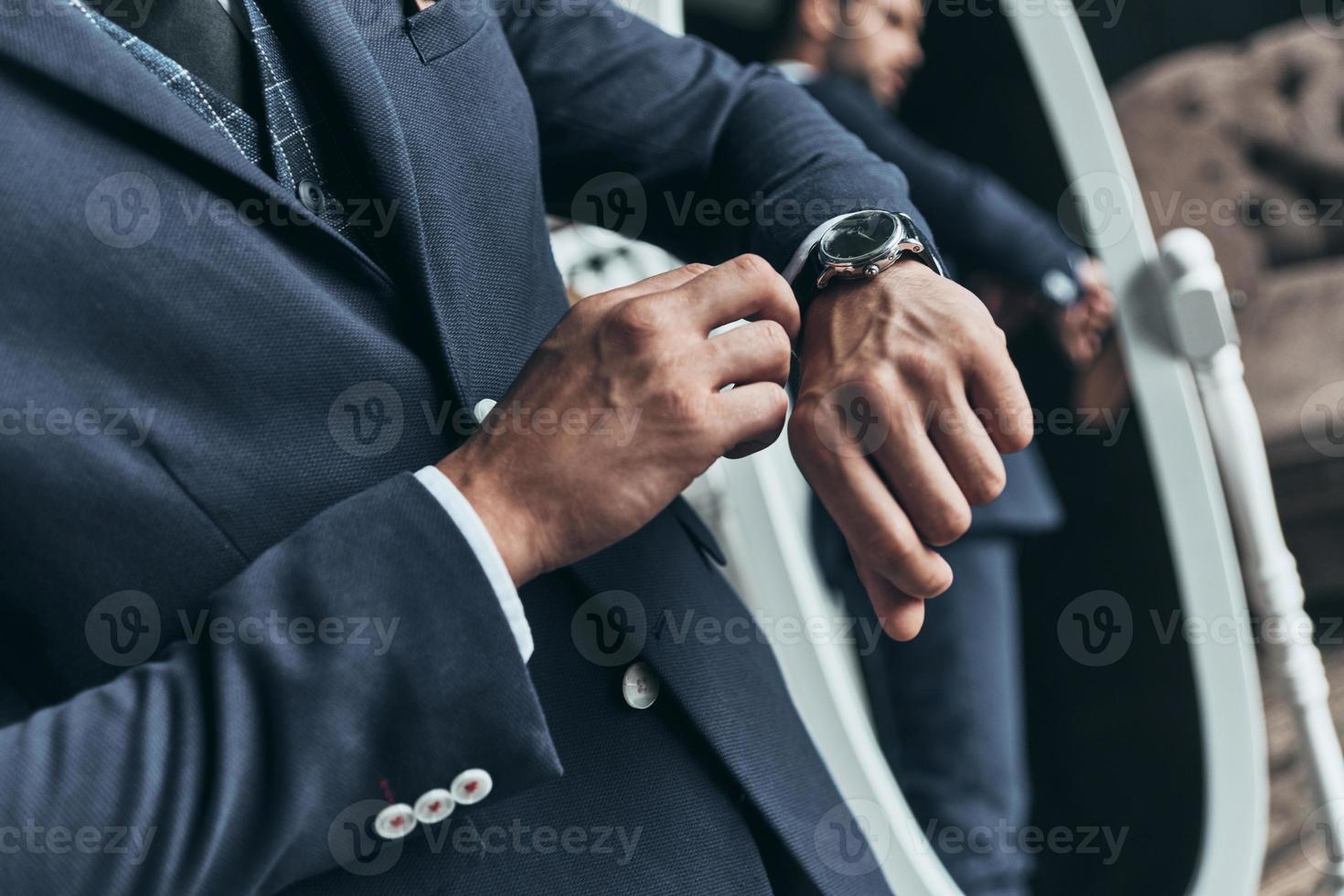 Always in time. Close up of young man in full suit checking the time while standing in front of the mirror indoors photo