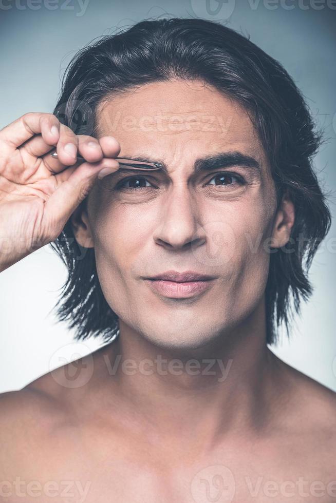 Tweezing eyebrows. Portrait of young shirtless man tweezing eyebrows and expressing negativity while standing against grey background photo