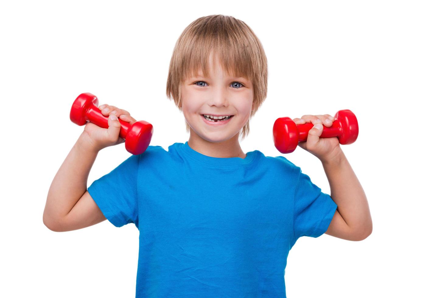 pequeño hombre fuerte. niño alegre mirando a la cámara y sonriendo mientras hace ejercicio con pesas y de pie aislado en blanco foto