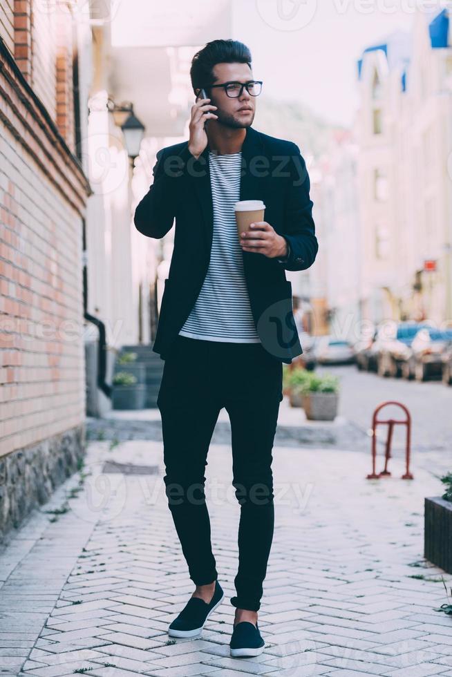 Talking with friend. Full length of handsome young man in smart casual wear carrying coffee cup and talking on mobile phone while walking along the street photo