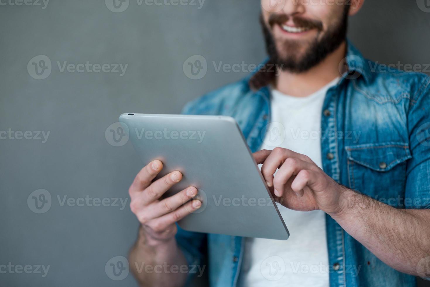 The future is in the touch. Cropped image of young man holding digital tablet and smiling while standing against grey background photo
