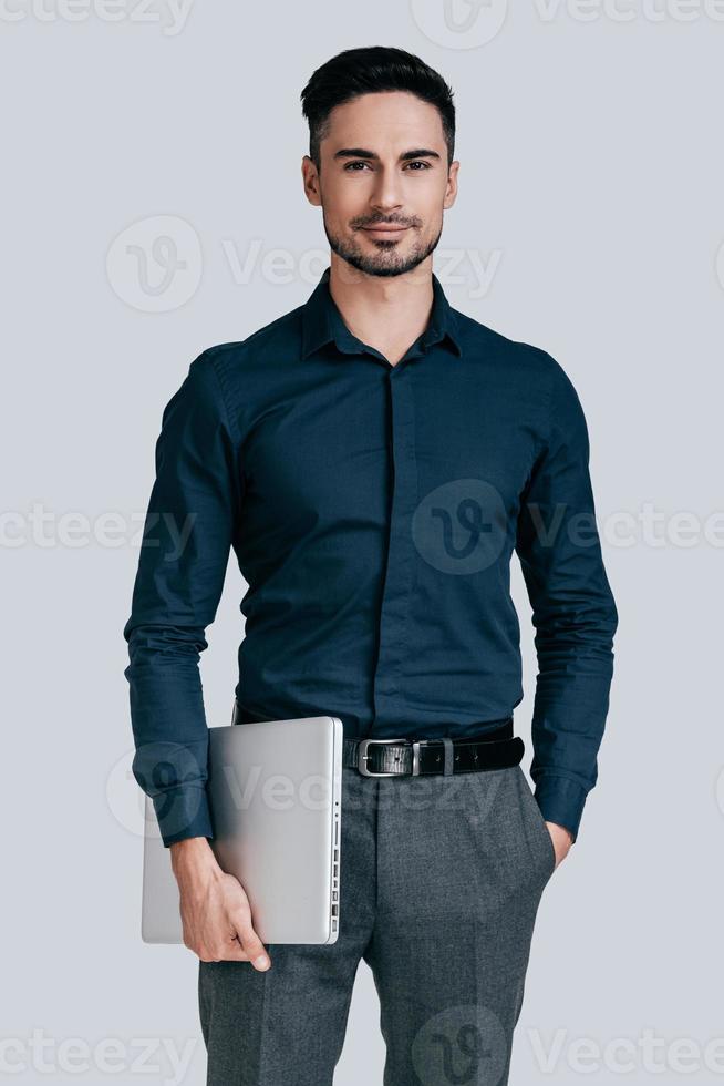 Confident businessman. Good looking young man in shirt carrying laptop and looking at camera with smile while standing against grey background photo