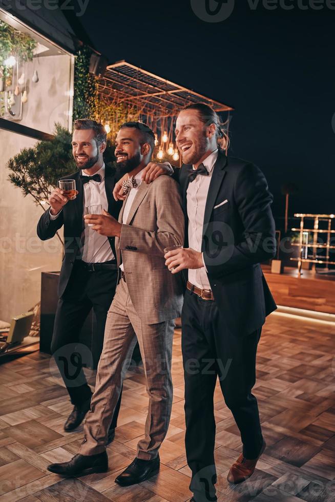 Full length of three handsome men in suits and bowties drinking whiskey and communicating photo