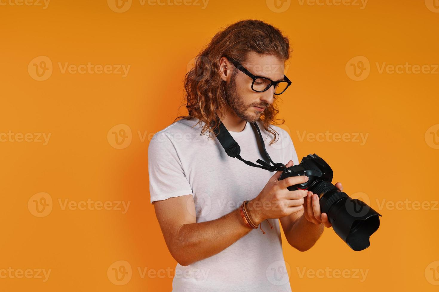 Charming young man in casual clothing using photographic camera while standing against yellow background photo