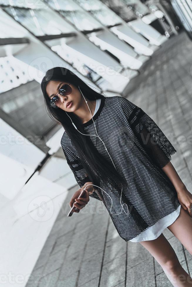 Music is her life. Beautiful young woman in casual wear holding a smart phone while standing outdoors photo