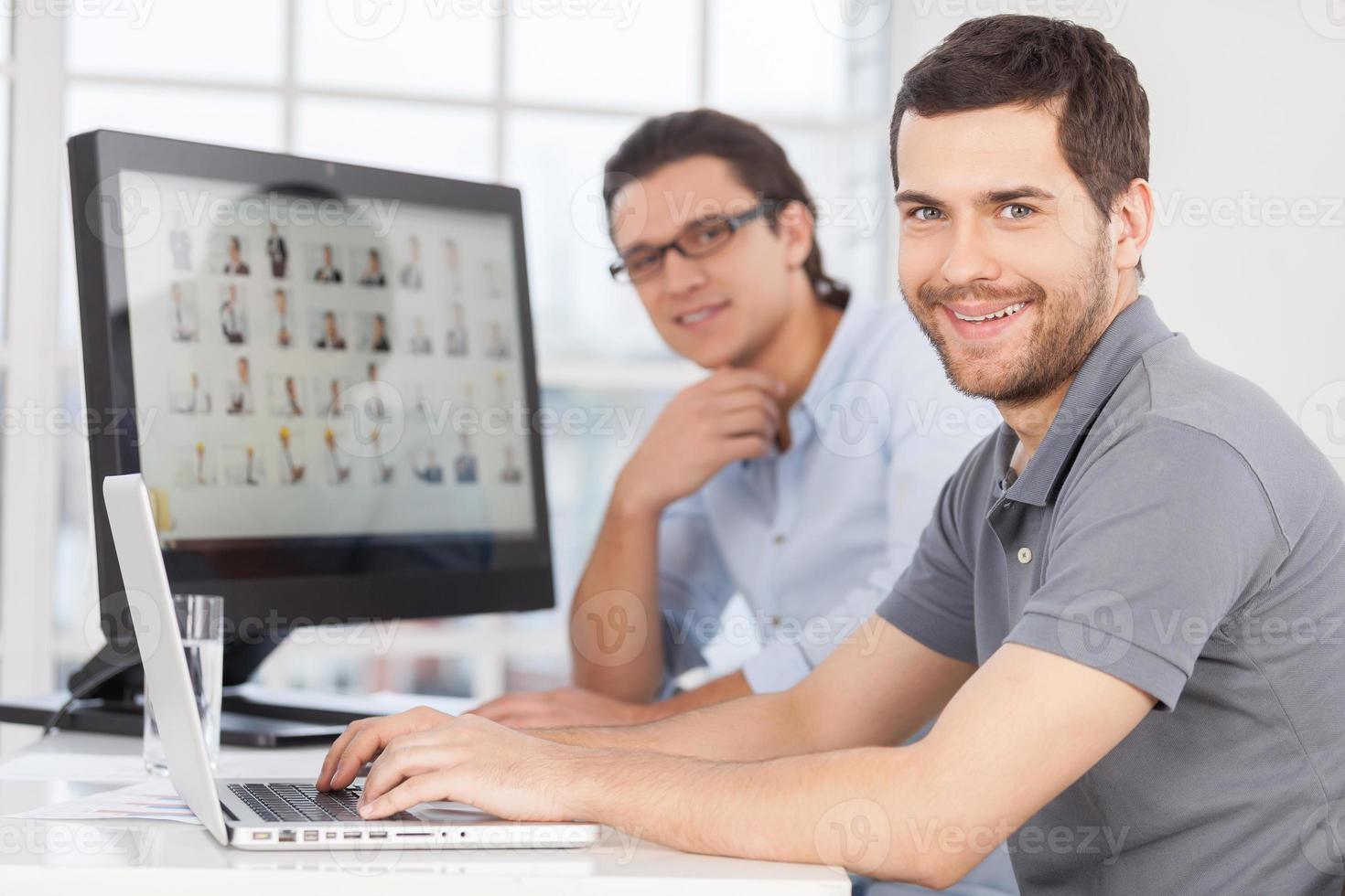 IT staff. Two cheerful young men smiling at camera while sitting in front of computer monitors photo