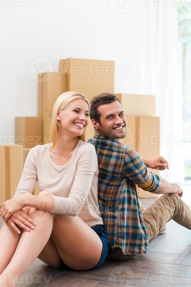 Starting new life together. Happy young couple sitting on the floor of their new apartment while cardboard boxes laying in the background photo