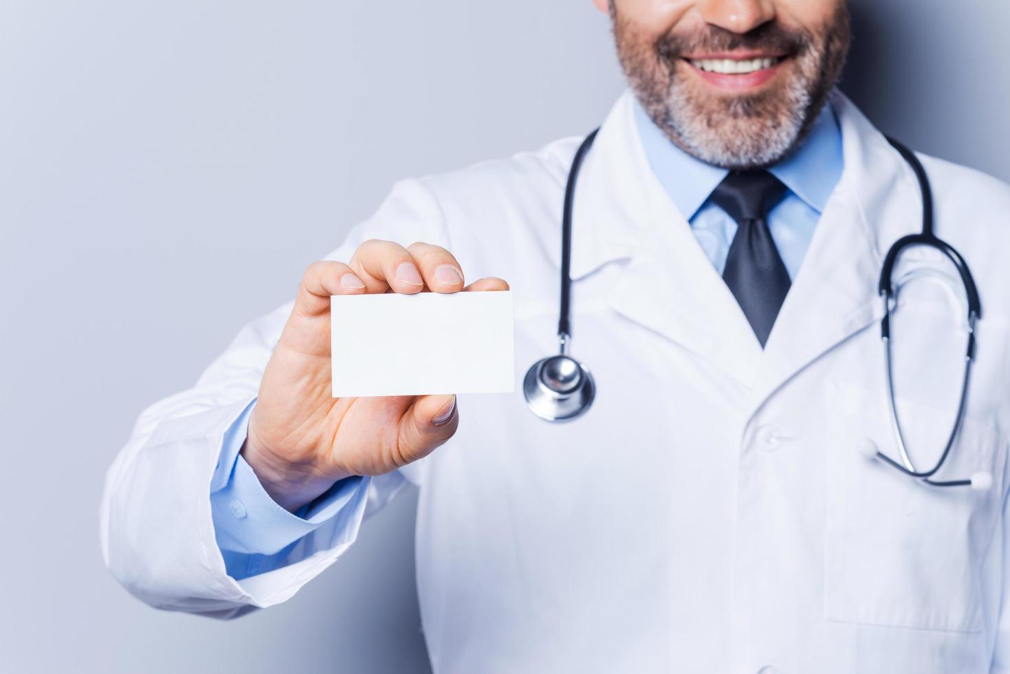 Doctor holding copy space. Close-up of confident mature doctor showing his business card and smiling while standing against grey background photo