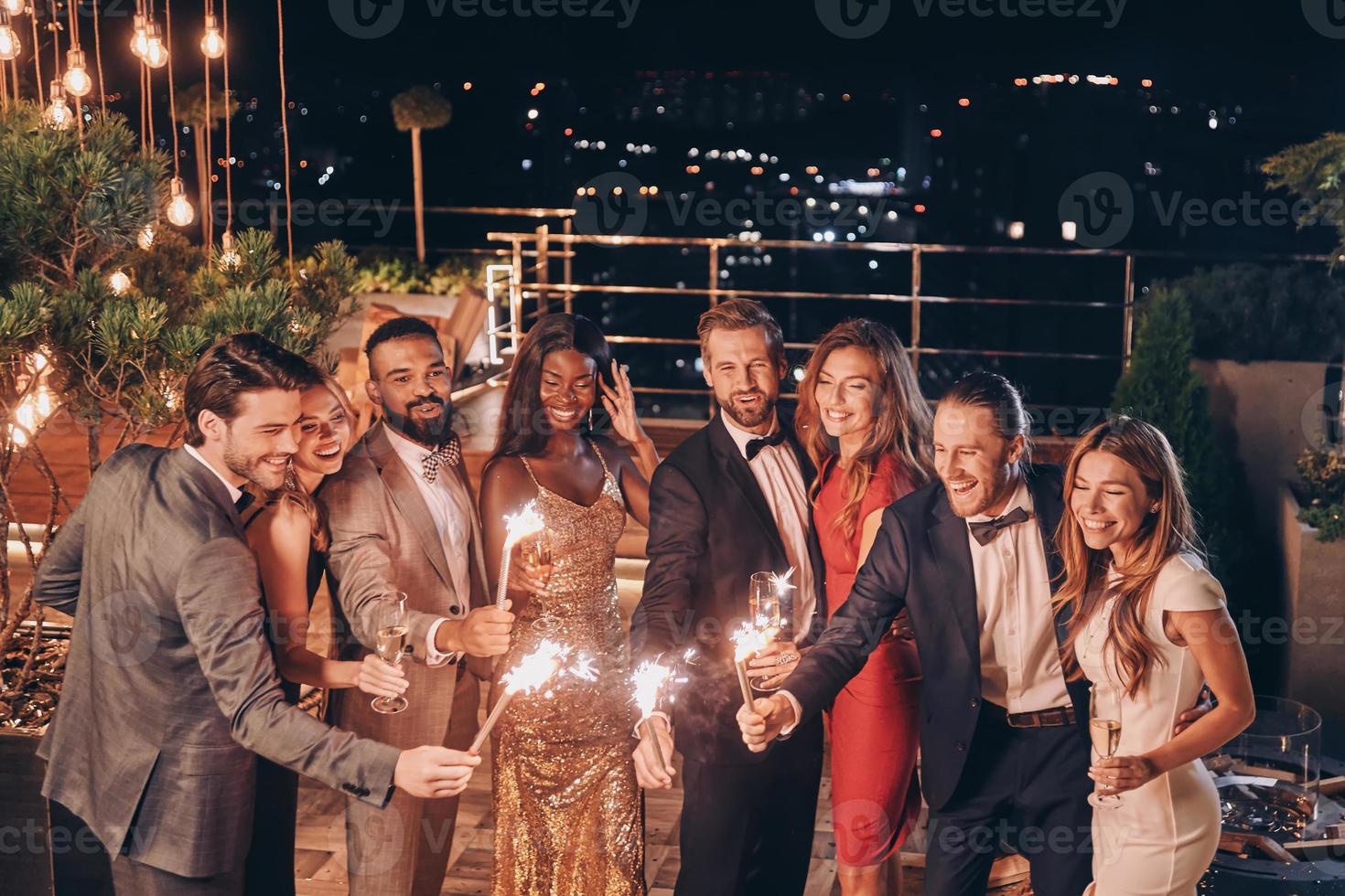 Group of beautiful people in formalwear holding sparklers and smiling while spending time on outdoor party photo