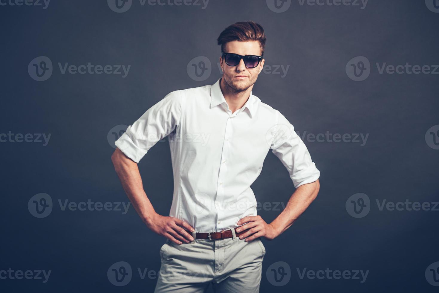 How you doing Confident young handsome man in sunglasses keeping arms akimbo and looking at camera while standing against grey background photo