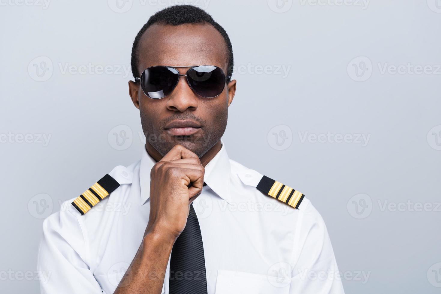 Confident and experienced pilot. Confident African pilot in uniform holding hand on chin while standing against grey background photo