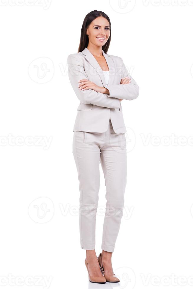Successful businesswoman. Full length of confident young businesswoman in suit keeping arms crossed and smiling while standing against white background photo
