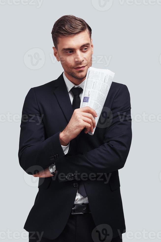 Too good to be real. Good looking young man in full suit holding newspaper and looking away while standing against grey background photo