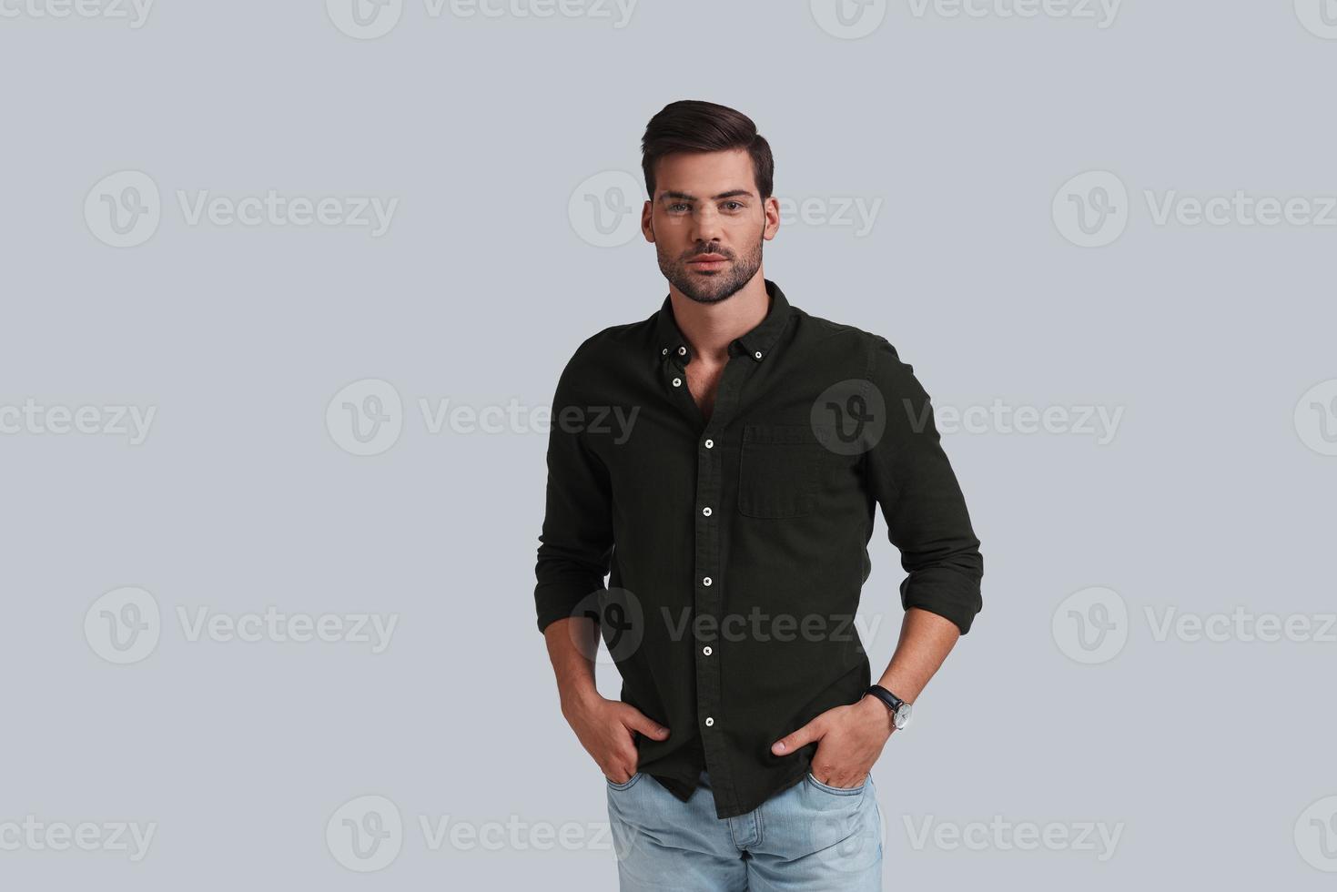 Confident man. Good looking young man keeping hands in pockets and looking at camera while standing against grey background photo