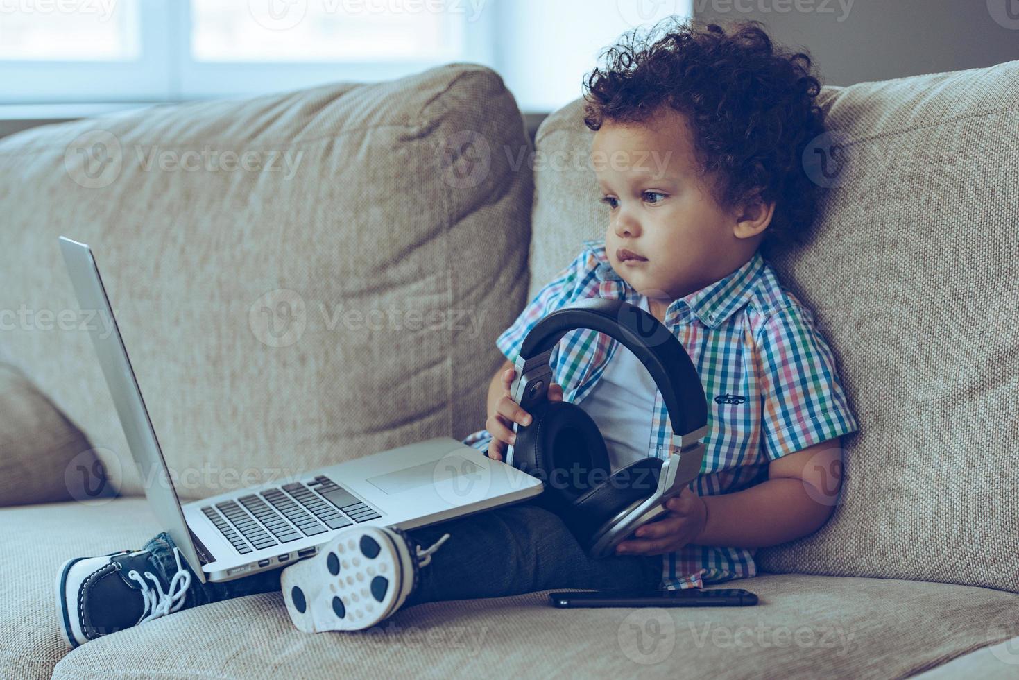 Technologies make life easier. Side view of little African baby boy looking at laptop and holding headphones while sitting on the couch at home photo