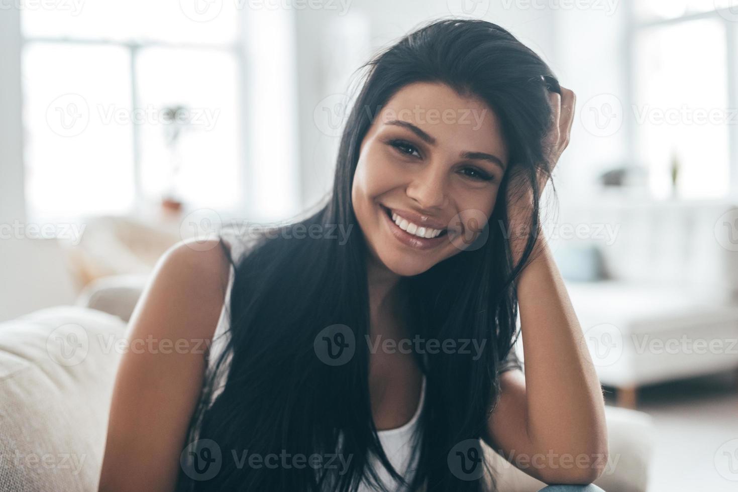 Perfect smile. Portrait of beautiful young woman looking at camera and smiling while sitting on the couch at home photo