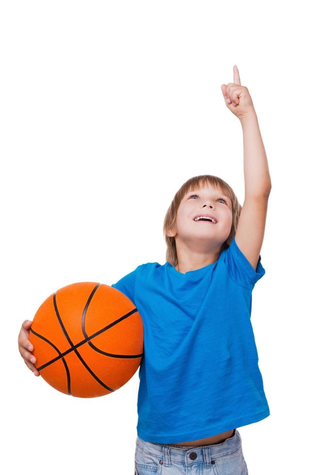 Curious little boy. Cheerful little boy holding basketball ball and pointing up while standing isolated on white photo