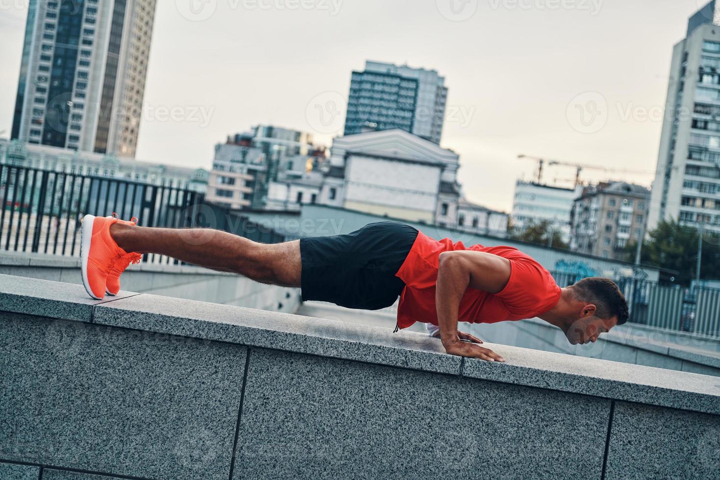 Confident young man in sports clothing keeping plank position while exercising outdoors photo