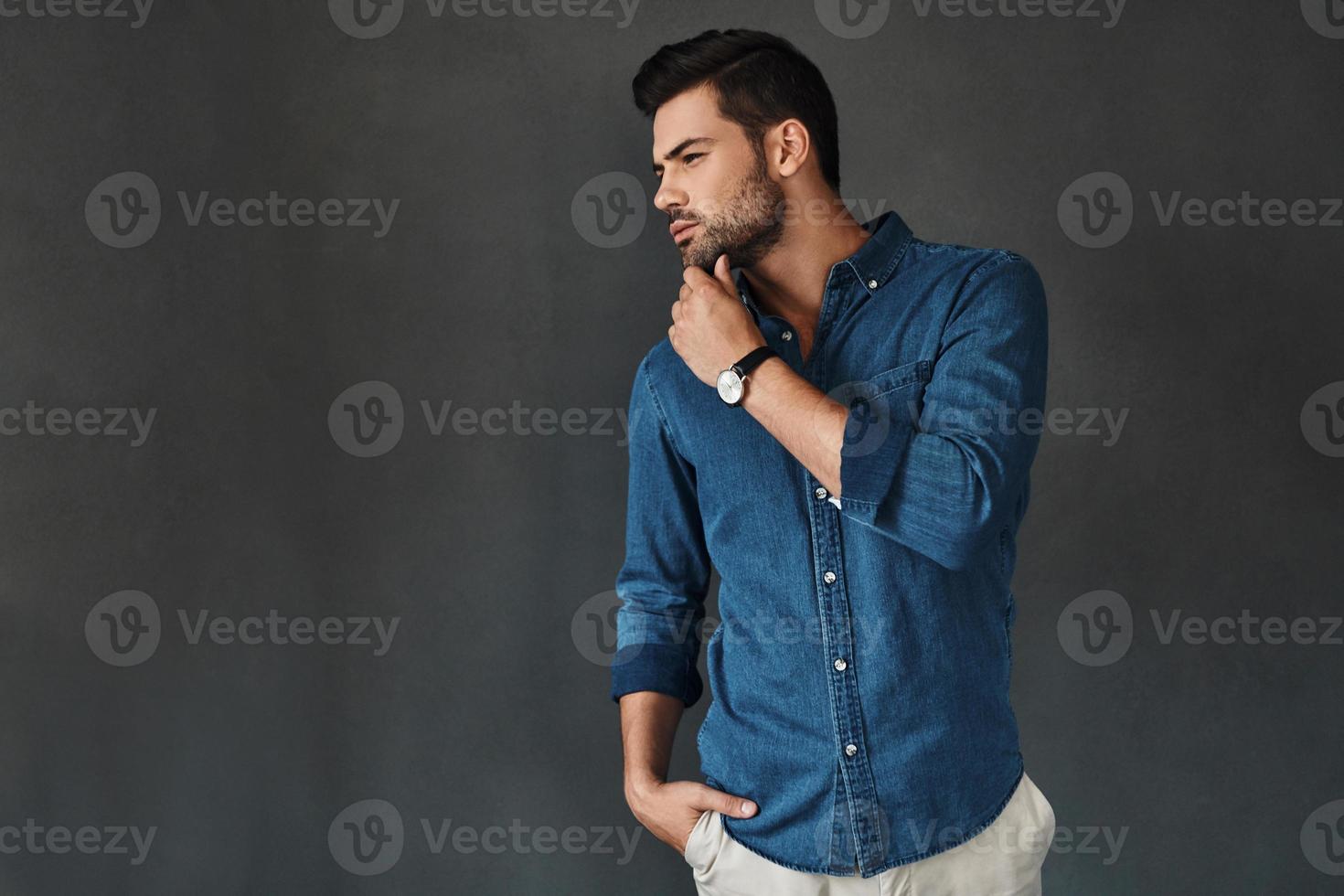 What if... Serious young man keeping hand on chin and looking away while standing against grey background photo