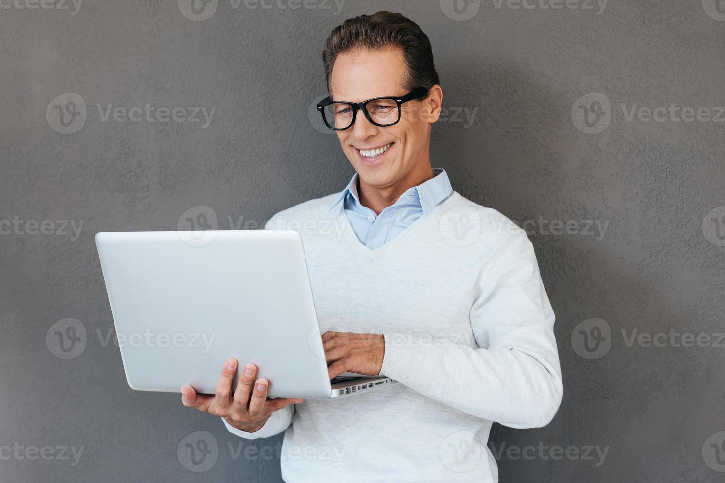 IT support. Confident mature man working on laptop and smiling while standing against grey background photo