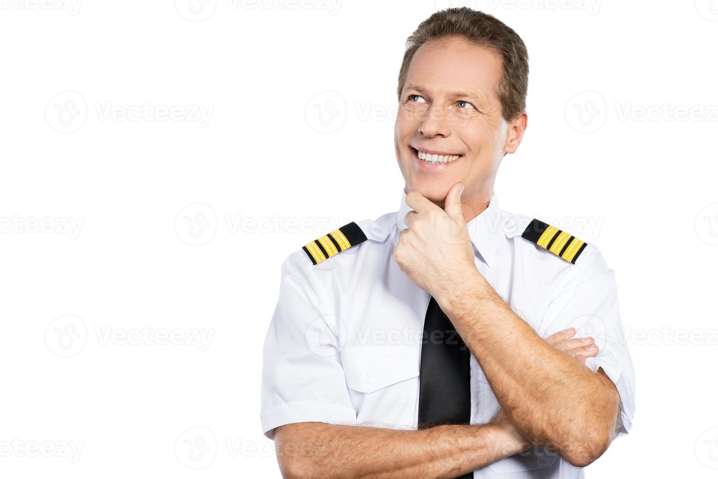 Passionate about sky. Thoughtful male pilot in uniform holding hand on chin and looking up with smile while standing against white background photo