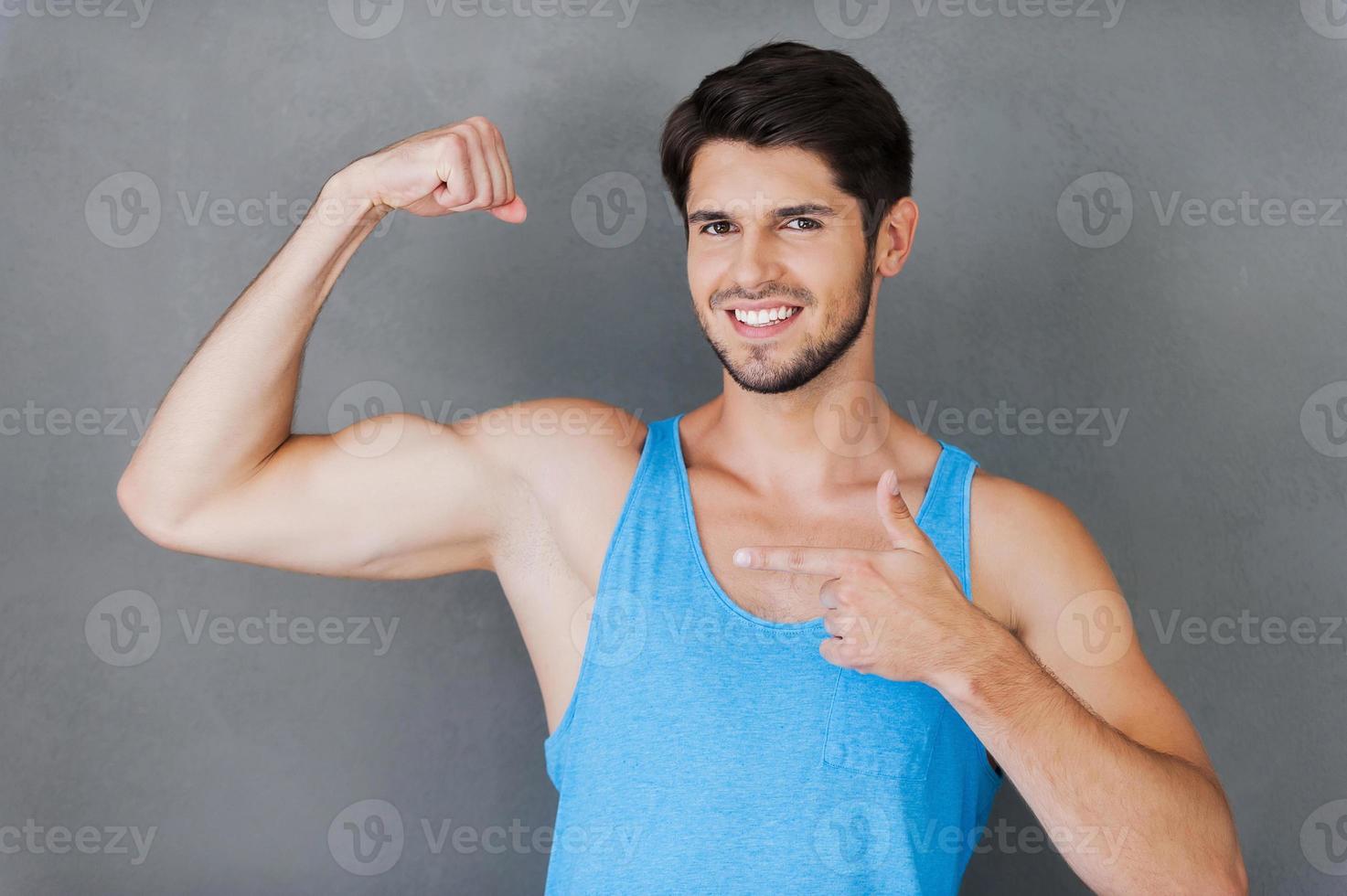 orgulloso de sus bíceps perfectos. alegre joven musculoso señalando su bíceps mientras está de pie contra el fondo gris foto