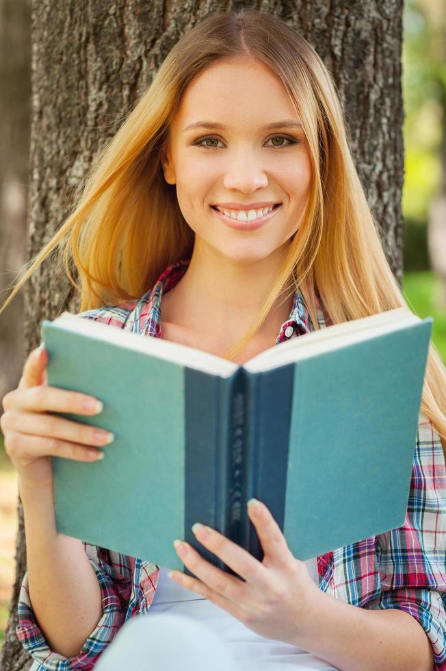 aire fresco y libro favorito. hermosa joven sosteniendo un libro y sonriendo mientras se apoya en el árbol en un parque foto