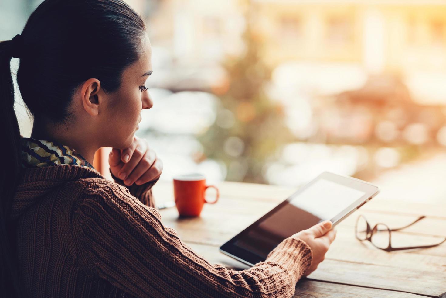 Enjoying work and fresh air. Side view of beautiful young woman holding digital tablet while sitting at the rough wooden table photo